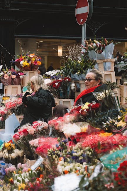 A busy flower market scene featuring a variety of fresh floral arrangements and bouquets. In the foreground, numerous vibrant bouquets display pink, red, white, and purple flowers wrapped in clear cel