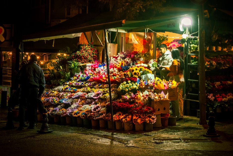 At a flower market stall on a street corner during nighttime, a vibrant display of fresh flowers in various colors is arranged in buckets and wrapped bouquets. The flowers include bright yellow sunflo