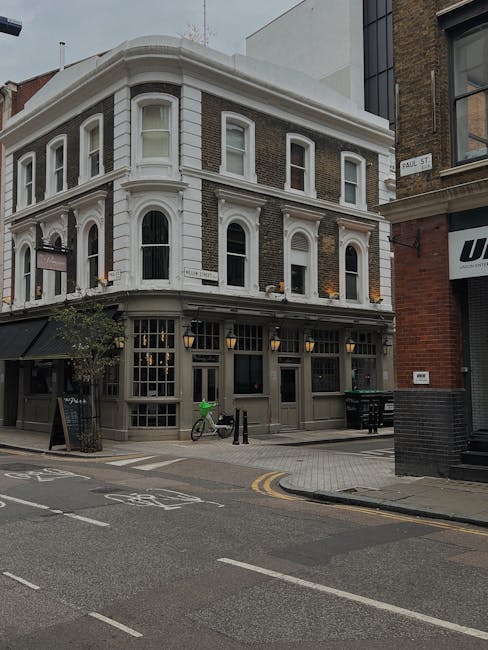 The image shows a corner building with a classic architectural style, featuring large bay windows and detailed white trim on the upper floors. The ground floor has a dark grey facade with large glass